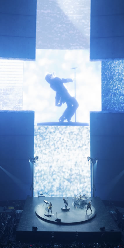 Bono sul palco ripreso da telecamera e proiettato su schermi alle spalle con una scenografia che disegna una croce